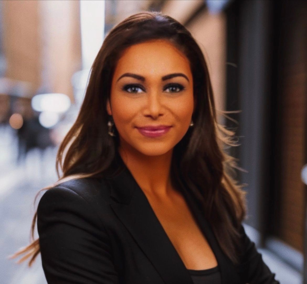 Woman with long hair and black blazer smiling at the camera, with a blurred street background.